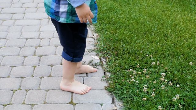 Slow motion footage of barefoot little boy standing on paved road and trying to step on fresh green grass at park