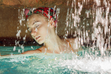 Portrait of a beautiful blonde young woman captured in swimming suit in a secret water cave. Her body is in the water, in front of her is waterfall and she enjoys the sun's rays on her soft skin.