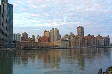 Fototapeta na wymiar Urban landscape at sunset in winter along East River in Manhattan, New York. Buildings reflection in harmony with pastel hues of cumulus clouds during the quiet city sunset.