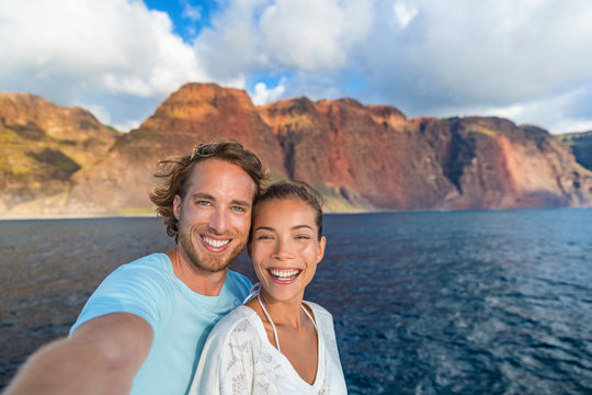 Happy couple tourists on sunset cruise in Na Pali Coast Kauai, Hawaii taking selfie photo with mobile phone app. Smiling Asian woman and Caucasian man taking picture on summer holiday.