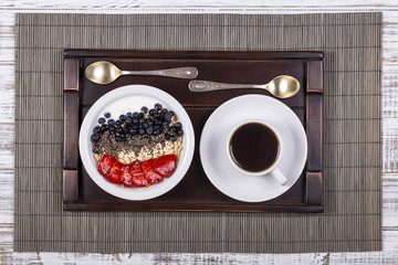 White cup of coffee and healthy muesli made from strawberry, blueberry, chia seeds, oat flakes with yogurt on a tray on white wooden table. Lifestyle concept. Top view