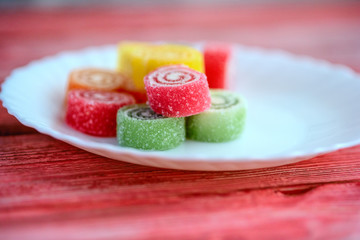 Multicolored marmalade on a white plate on a light red wooden background