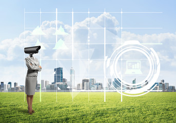 Camera headed woman standing on green grass against modern cityscape