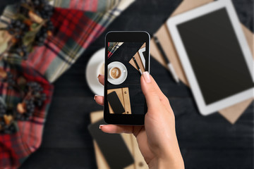 Woman taking picture of coffee on her smartphone. Top view of the table: laptop, smartphone, woman hands, notebook.