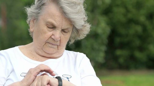 Black wristband. Mature old woman dressed in white t-shirt looks at the results of physical activity using a wristband fitness tracker. Positive person. Excellent mood