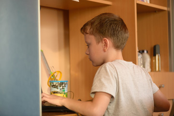 The boy prints the text on the computer keyboard in his room.Portrait of a boy in the children's room with a computer at his workplace. Sits at a table by the cupboard with shelves.