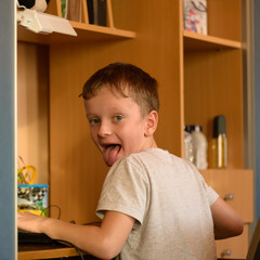 Naklejka premium The boy prints the text on the computer keyboard in his room.Portrait of a boy in the children's room with a computer at his workplace. Sits at a table by the cupboard with shelves.
