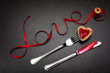 Inscription, word love of red satin ribbon and heart of toast bread with red jam on fork and knife with jam.Valentine day background.Love concept.On dark stone background.Creative.Love backgound