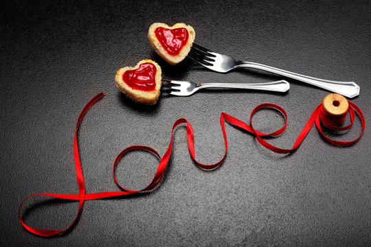 Inscription, word love of red satin ribbon and two hearts of toast bread with red jam on fork.Valentine day background.Love concept.On dark stone background.Creative.Love background.