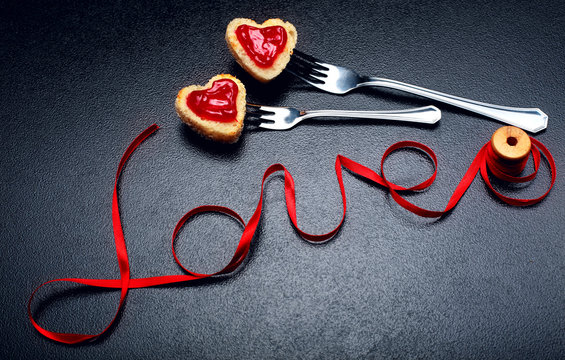 Inscription, word love of red satin ribbon and two hearts of toast bread with red jam on fork.Valentine day background.Love concept.On dark stone background.Creative.Love background.