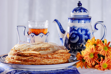 Freshly baked pancakes, a teapot, a glass with tea, sour cream are on a table set for the maslenitsa, pancake week celebration