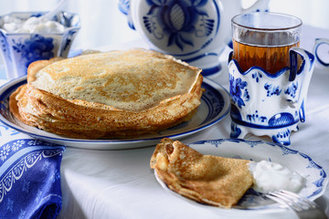Freshly baked pancakes, a teapot, a glass with tea, sour cream are on a table set for the...
