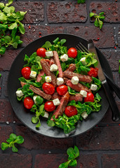 Juicy Beef Sirloin Steak Salad with roasted tomatoes, feta cheese and green vegetables in a black plate. healthy food