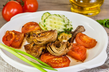 Half of an artichoke on the grill with slices of cucumber and ripe red tomato on a white plate with Brussels sprouts and green onions on the napkin with the devices.