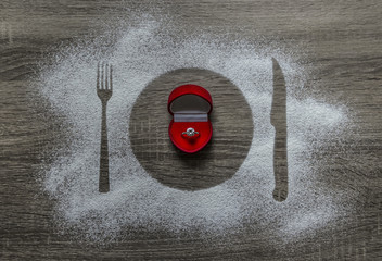 on a wooden background with powdered powder snow covered with a silhouette a plate a fork knife of appliances in the center lies a box with a ring with a diamond 