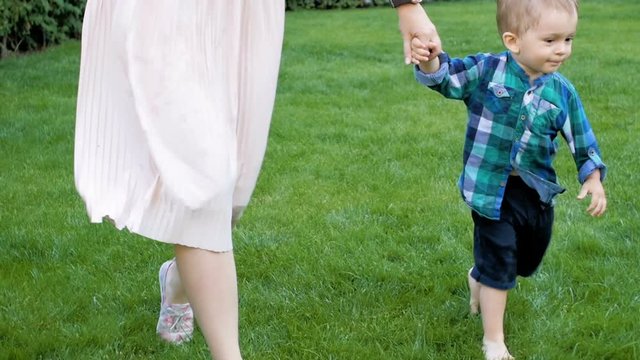 Slow motion video of adorable barefoot toddler boy holding mothers hand and walking on grass at park