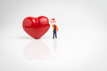 Miniature people, man holding sign with text I love you asking for love with shiny red heart on seamless white background