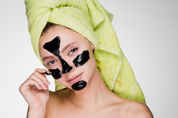 lovely girl with a green towel on her head apply a black mask against acne on problem areas on the...