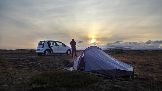 Mann steht zwischen Zelt und Auto und beotrachtet Sonnenuntergang - Island