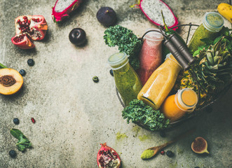 Colorful smoothies in bottles with fresh fruit and greens in basket on grey concrete background,...