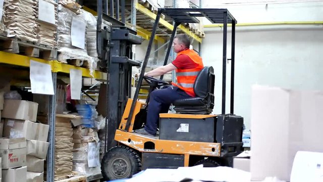 Forklift pallet with boxes in large modern warehouse. Warehouse worker driver in uniform moving cardboard boxes by forklift stacker loader