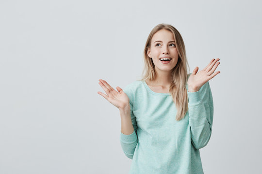 Indoor shot of excited blonde beautiful young woman with long hair looks with great surprisment at camera, recieves news from best friend isolated against gray backgroud. Positive emotions concept.