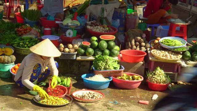 The woman at a counter of vegetables and fruit touches small pepper. The woman squats in the Vietnamese hat. The market in Vietnam. Vietnam