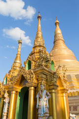 Fototapeta premium Two small pagodas and statues in front of the gilded Shwedagon Pagoda in Yangon, Myanmar on a sunny day.