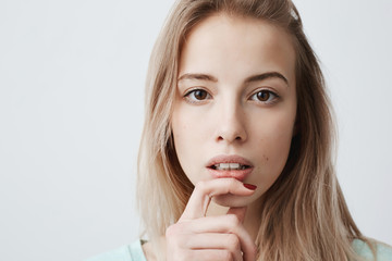 Naklejka premium European beautiful young female with blonde dyed straight hair looks at camera with interest, has pensive expression of face, makes decision, holds finger on chin, isolated against gray background.