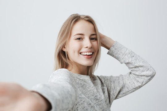 Attractive dark-eyed blonde female dressed casually having delightful look smiling broadly. Beautiful woman having cheerful expression while posing against gray background, stretching arm to camera.