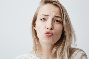 Cute young female with pale skin, long blonde hair, wears casual sweater, rounds lips, looks coquette, isolated against gray background. Fashionable stylish woman sends kisses