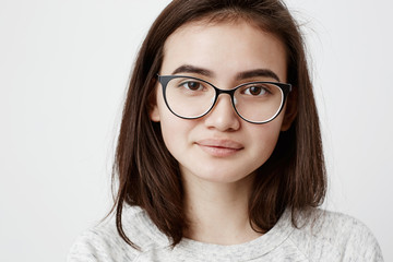 Close-up portrait of stylish dark-haired beautiful teenage girl with trendy haircut dressed in t-shirt, looks at camera, wears trendy eyewear, smiles gently. People lifestyle youth and beauty
