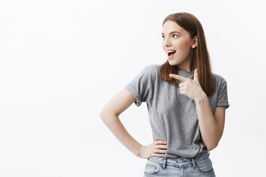 Copy space. Good-looking cinfident young european student with dark hair in casual grey clothes looking aside with opened mouth and excited expression, pointing with finger on free space on white