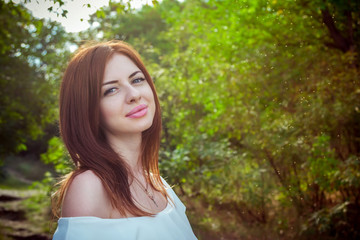 Portrait of beautiful young smiling woman with long red hair, wearing elegant white top in a forest with green trees on a sunny summer day