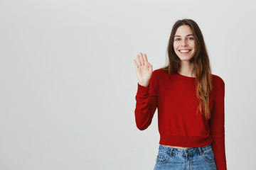 Hi there. Positive emotions. Copy space. Young good-looking caucasian girl with brown long hair in red sweater and denim jeans smiling, waving hand with relaxed and happy face expression.