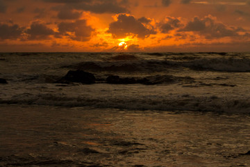 Obraz premium Waves crash ashore as the Southern Pacific sun sets in the background, turning the sky to liquid orange fire that is reflected in the water.