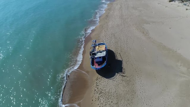 Aerial flying around stranded refugee boat on empty mediterranean beach showing azure blue ocean waves and peaceful beach with human footprints around the stranded ship taken during refugee crisis 4k