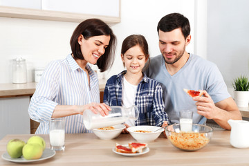 Happy family have healthy breakfast together. Smiling mother pours milk in bowl with cornflakes, eat apples, snacks and drink milk, sit together at kitchen. Healthy food, family and eating concept.