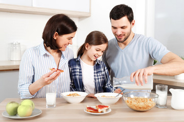 Happy young parents and their lovely daughter sit together at kitchen table, eat flakes, have healthy breakfast, enjoy good morning, have friendly relationship. Family and eatting concept.