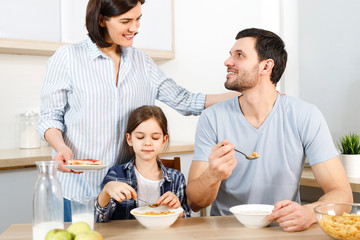 Obraz na płótnie Canvas Three family members have delicious healthy breakfast at kitchen, eat cornflakes with milk, enjoy togetherness and domestic atmosphere. Husband looks with love at wife, satisfied with tasty dish.