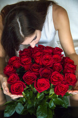 Brown hair girl is smelling a bouquet of red roses, holding them in hands and siting on white sofa