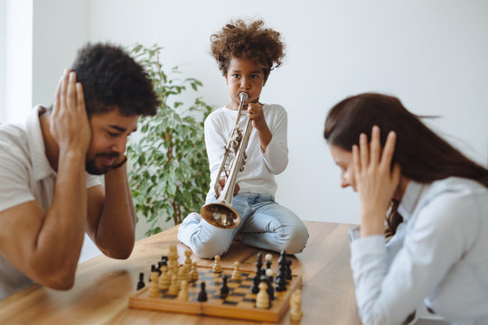 Mother and father trying to play chess while their child plays trumpet