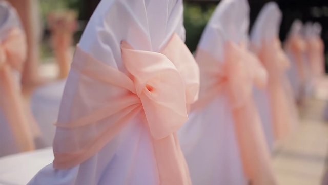 Wedding decor on the chairs. White wedding chairs with pink ribbon bow. Outdoors wedding ceremony. Rows of white wooden empty chairs on lawn before wedding ceremony. 