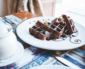 a beautifully decorated wooden table by the window with pastries and a waffle with berry sauce or chocolate with a cup of tea