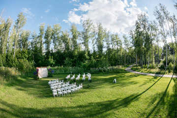 Beautiful setting for outdoors wedding ceremony waiting for bride and groom and guests. White chairs decorated with flowers, are in the zone of the wedding ceremony in a forest
