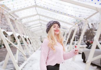 Outdoor portrait of positive young woman with bright makeup, wearing trendy knitted hat and sweater, posing at the city in winter