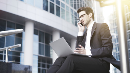 A handsome young businessman wearing glasses and a suit sitting on the stairs and working with laptop. Downtown.