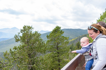 Fototapeta na wymiar Young woman hiking with baby in backpack looking at sweeping mountain views at scenic overlook