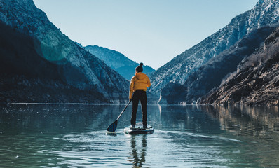 a woman with yellow plumber paddle surfing on a lake in a snowy mountain landscape - obrazy, fototapety, plakaty