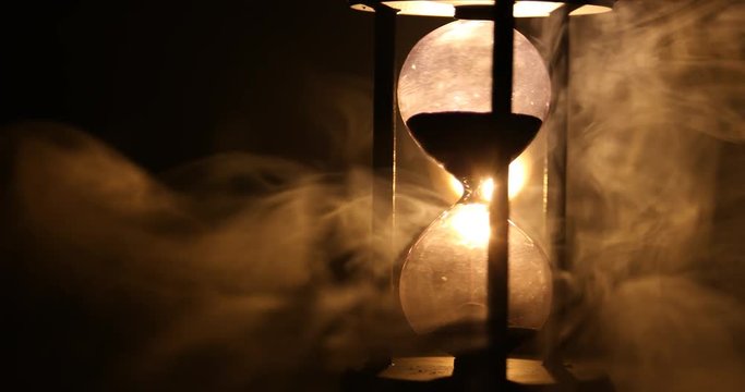 Time concept. Silhouette of Hourglass clock rotating in smoke on dark toned background, or symbols of time under smoke, the end
